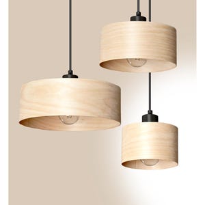 May include: A cluster of three pendant lights featuring cylindrical light-coloured wood veneer shades. Each shade is suspended by a thin black cord attached to a black metal fixture, with a single exposed light bulb centred inside each unit.