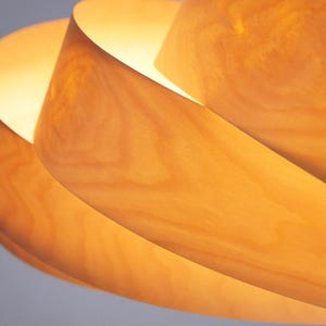 Op de afbeelding: Close-up van een houten hanglamp. De lamp heeft een spiraalvormig ontwerp met overlappende houten lamellen, waardoor een warme gloed ontstaat. De houtnerf is zichtbaar en voegt textuur toe. Het licht is warmgeel.