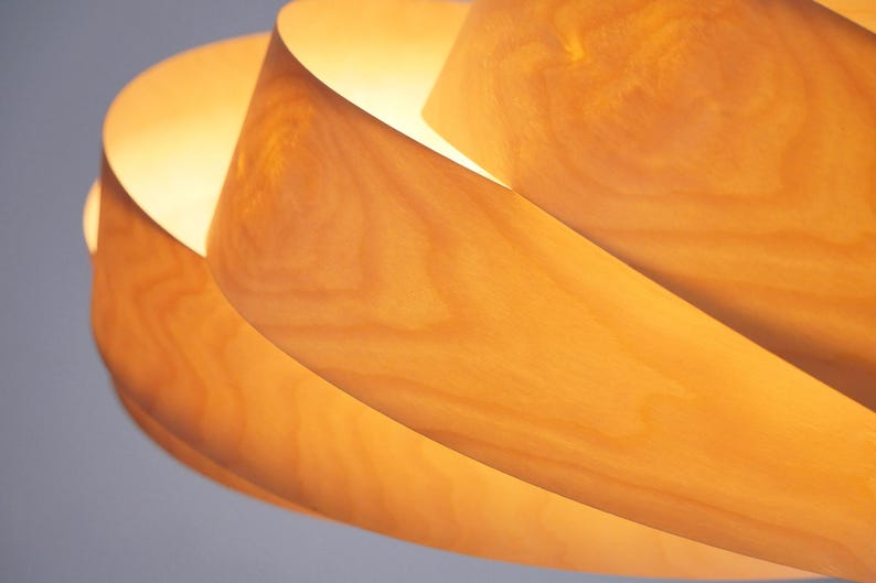 Op de afbeelding: Close-up van een houten hanglamp. De lamp heeft een spiraalvormig ontwerp met overlappende houten lamellen, waardoor een warme gloed ontstaat. De houtnerf is zichtbaar en voegt textuur toe. Het licht is warmgeel.