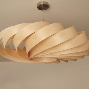 Op de afbeelding: Een moderne hanglamp van licht hout. De armatuur hangt aan het plafond en heeft een spiraalvormig ontwerp gemaakt van gelaagde houten stroken. Het licht heeft een natuurlijke houttint en een plafondbevestiging van geborsteld metaal.