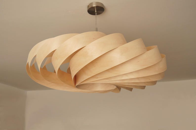 Op de afbeelding: Een moderne hanglamp van licht hout. De armatuur hangt aan het plafond en heeft een spiraalvormig ontwerp gemaakt van gelaagde houten stroken. Het licht heeft een natuurlijke houttint en een plafondbevestiging van geborsteld metaal.