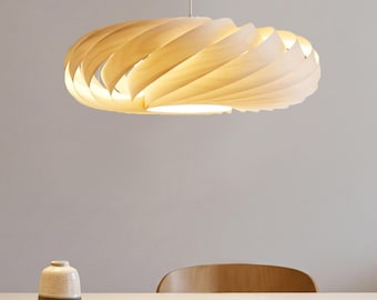 Może przedstawiać: Lampa wisząca z okrągłym, spiralnym kloszem wykonanym z jasnych drewnianych listewek, które emitują ciepłe, rozproszone światło. Oprawa wisi na cienkim przewodzie nad stołem, na którym stoi mały, dwukolorowy wazon ceramiczny.
