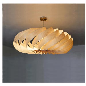 Può includere: Una moderna lampada a sospensione in legno. La lampada ha un design a spirale realizzato con doghe di legno curve e stratificate. L'apparecchio è appeso a un supporto a soffitto e un filo color argento. La luce è accesa, illuminando l'interno.