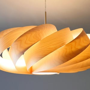 Op de afbeelding: Een moderne houten hanglamp. De lamp heeft een spiraalvormig ontwerp gemaakt van gebogen, lichtgekleurd hout. De lamp hangt aan het plafond met een dun snoer. Het licht geeft een warme, uitnodigende gloed, perfect voor de inrichting van een huis.