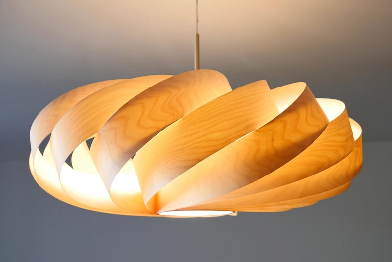 Op de afbeelding: Een moderne houten hanglamp. De lamp heeft een spiraalvormig ontwerp gemaakt van gebogen, lichtgekleurd hout. De lamp hangt aan het plafond met een dun snoer. Het licht geeft een warme, uitnodigende gloed, perfect voor de inrichting van een huis.
