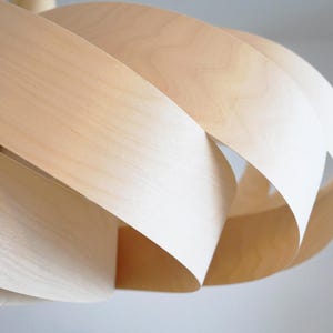Op de afbeelding: Een moderne hanglamp van lichtgekleurd hout. Het ontwerp bestaat uit gelaagde, gebogen houten stroken, die een open, sculpturale vorm cre&euml;ren. De natuurlijke houttinten en de vorm van de lamp suggereren een moderne esthetiek.