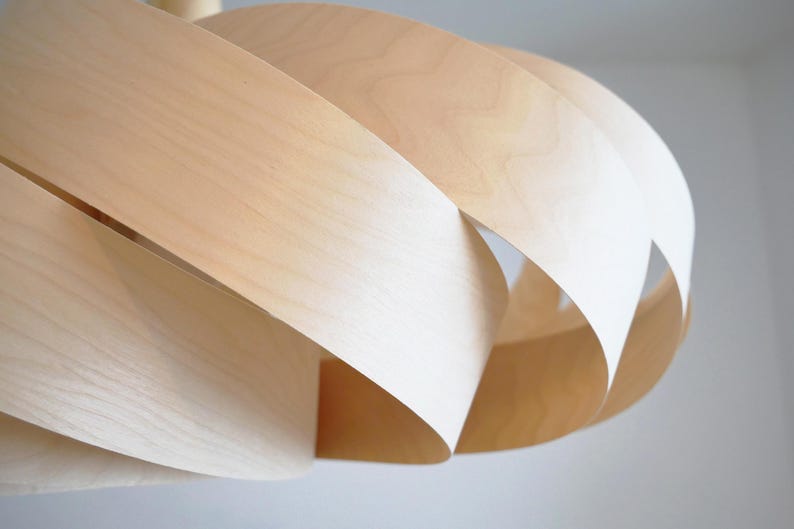 Op de afbeelding: Een moderne hanglamp van lichtgekleurd hout. Het ontwerp bestaat uit gelaagde, gebogen houten stroken, die een open, sculpturale vorm cre&euml;ren. De natuurlijke houttinten en de vorm van de lamp suggereren een moderne esthetiek.