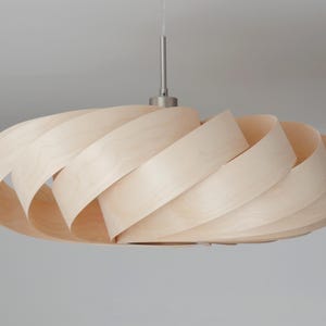 Op de afbeelding: Een moderne hanglamp van lichtgekleurd hout. De lampenkap heeft een spiraalvormig ontwerp gemaakt van overlappende houten stroken. Hij hangt aan een zilverkleurige metalen staaf.