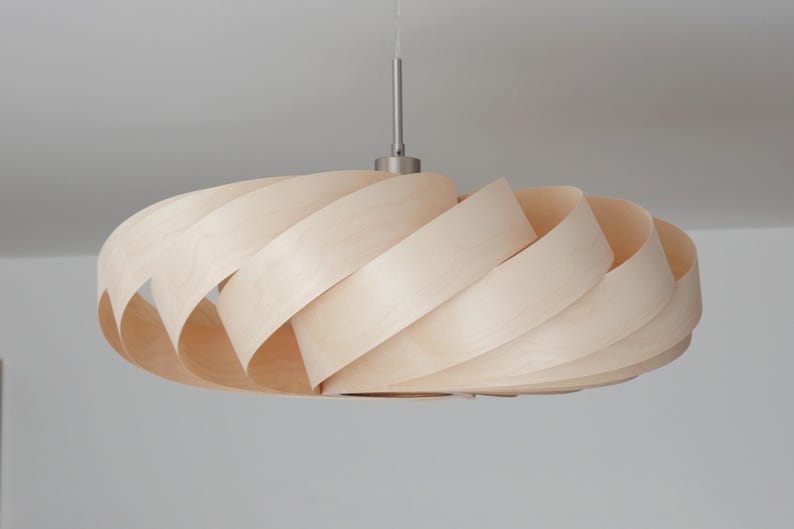 Op de afbeelding: Een moderne hanglamp van lichtgekleurd hout. De lampenkap heeft een spiraalvormig ontwerp gemaakt van overlappende houten stroken. Hij hangt aan een zilverkleurige metalen staaf.