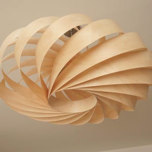 Op de afbeelding: Een lichtgekleurde houten hanglamp. De lamp bestaat uit talrijke gebogen houten stroken die in een spiraal zijn gerangschikt, waardoor een open, luchtig ontwerp ontstaat. De lamp hangt aan het plafond en toont een moderne, natuurlijke esthetiek.