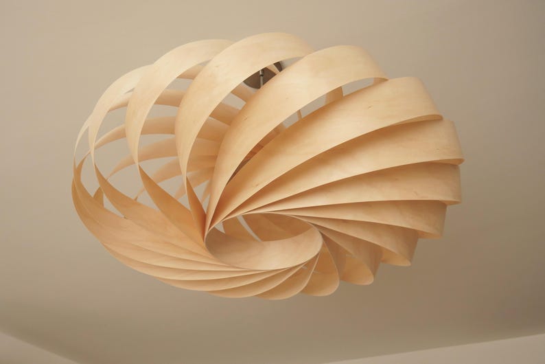 Op de afbeelding: Een lichtgekleurde houten hanglamp. De lamp bestaat uit talrijke gebogen houten stroken die in een spiraal zijn gerangschikt, waardoor een open, luchtig ontwerp ontstaat. De lamp hangt aan het plafond en toont een moderne, natuurlijke esthetiek.
