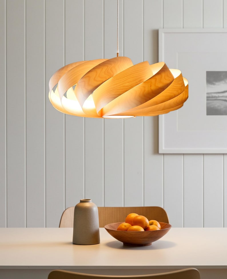 May include: A wooden pendant light with a layered, swirling design, suspended from the ceiling. The light emits a warm glow. The fixture is made of light-coloured wood. The setting is a dining room.