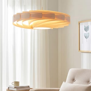 Pode incluir: Uma luminária pendente circular com várias ripas de madeira clara sobrepostas em um padrão espiral. A lâmpada emite um brilho quente e difuso a partir do centro, suspensa por um fio branco fino preso ao teto.