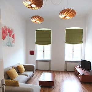 Op de afbeelding: Een moderne woonkamer met drie houten spiraalvormige hanglampen. De kamer heeft een lichtgrijze hoekbank met gele kussens, een houten salontafel en een tv-meubel. Twee ramen hebben groene jaloezie&euml;n en een schilderij van rode bloemen hangt aan de muur.
