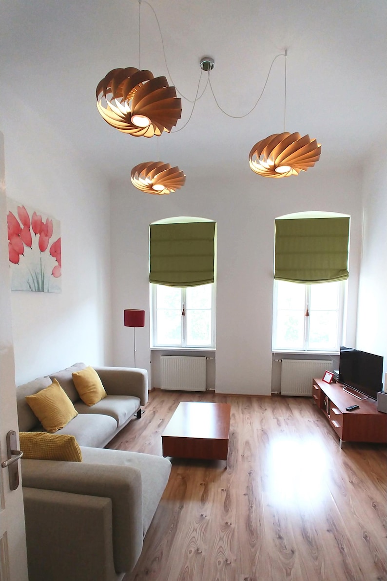 Op de afbeelding: Een moderne woonkamer met drie houten spiraalvormige hanglampen. De kamer heeft een lichtgrijze hoekbank met gele kussens, een houten salontafel en een tv-meubel. Twee ramen hebben groene jaloezie&euml;n en een schilderij van rode bloemen hangt aan de muur.