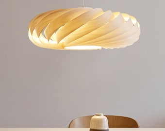Può includere: Una lampada a sospensione circolare caratterizzata da una serie di listelli in legno chiaro curvi e sovrapposti a spirale, che emette una luce calda dall'interno, sospesa tramite un cavo sottile dal soffitto sopra un tavolo.
