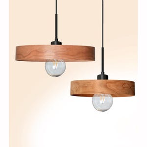 Op de afbeelding: Twee hanglampen met een minimalistisch design. Elke lamp heeft een cirkelvormige, lichtbruine houten kap, een heldere glazen lamp en een zwarte staaf. De lampen hangen tegen een crèmekleurige achtergrond.