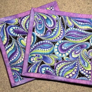 May include: Two purple and white quilted pot holders with a paisley pattern in shades of blue, green, and white.