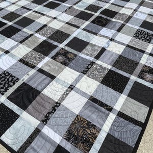 May include: A patchwork quilt featuring a grid of black, white, and gray fabric squares. The design incorporates various patterns including solid colors, dots, swirls, and firework motifs, all joined by white sashing and finished with black binding.