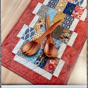 May include: A rectangular quilted table runner featuring a patchwork of floral and geometric patterns in blue, orange, yellow, and white, bordered by a solid coral fabric. Two wooden salad servers are crossed on top. Text reads CHA CHA CHA!