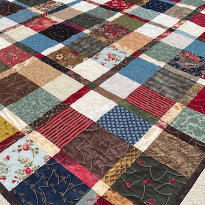 May include: A patchwork quilt featuring a grid of square fabric blocks in various colors including red, blue, green, brown, and cream. The blocks display diverse patterns such as floral prints, stripes, and textures, all joined by visible stitching.