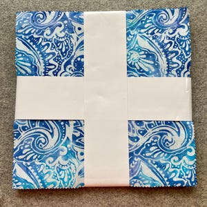 May include: A stack of square fabric pieces featuring a blue and white paisley pattern with pinked edges. The fabric squares are held together by a white paper band wrapped in a cross shape around the center of the stack, securing the collection.