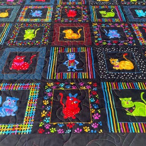 May include: A colorful quilt featuring a repeating pattern of cartoon cats in various colors, each framed in a square. The squares are set against a black background with rainbow borders and paw prints. The quilt is machine-stitched.