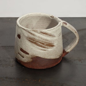 May include: Hand-thrown ceramic coffee mug with matte cream upper glaze and rich, earthy brown base. Features organic brushstroke patterns, a gently tapered profile, and a sturdy curved handle. Its artisanal texture and warm tones evoke a cozy, rustic farmhouse aesthetic.