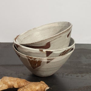 Peut inclure: Trois bols en céramique empilés sur une surface sombre. Les bols sont gris clair avec des motifs abstraits bruns. Deux morceaux de gingembre frais sont au premier plan. Les bols sont probablement destinés à servir des aliments.