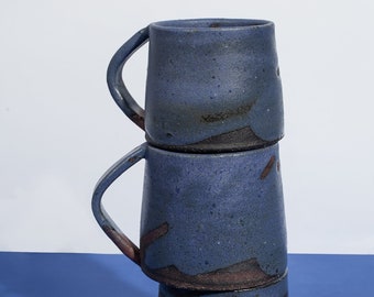 Può includere: Tre tazze in ceramica impilate con una glassa blu scuro e accenti marrone scuro. Ogni tazza ha un manico curvo e una forma leggermente affusolata. Le tazze sono disposte su una superficie blu su sfondo bianco.