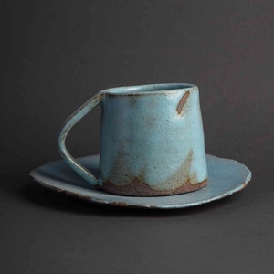 Peut inclure: Un ensemble de tasse et soucoupe en céramique bleu clair avec un glaçage rustique et irrégulier. La tasse a une poignée arrondie et la soucoupe a une forme légèrement irrégulière.