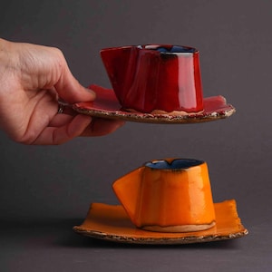 Può includere: Due tazze da caffè e piattini in ceramica. Una è rossa con interno blu, l'altra arancione con interno blu. Le tazze hanno un design angolare unico e poggiano su piattini rettangolari abbinati.
