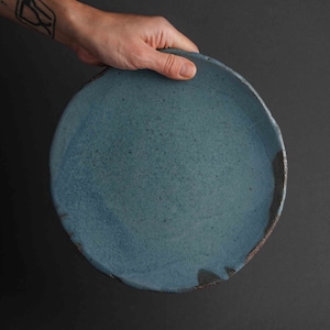 May include: A large, round, blue ceramic plate with a slightly uneven rim. The plate has a speckled, textured surface.