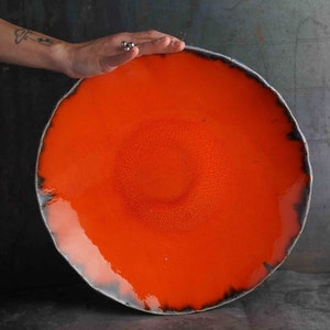 Op de afbeelding: Grote, ronde, oranje keramische serveerschaal met een getextureerd oppervlak. De rand is donkergrijs met een witte rand. De schaal is handgemaakt en heeft een glanzende afwerking. Een hand rust op de schaal.