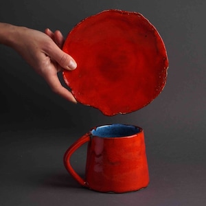 May include: A red ceramic mug with a blue interior and matching plate. The mug has a handle and a slightly irregular shape. The plate is round with a rough edge. The items are displayed against a dark gray background.
