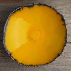 May include: A handmade, round ceramic bowl with a vibrant yellow interior and a dark blue, textured rim. The bowl's surface reflects light, and it sits on a wooden surface. This decorative bowl is ideal for serving food.