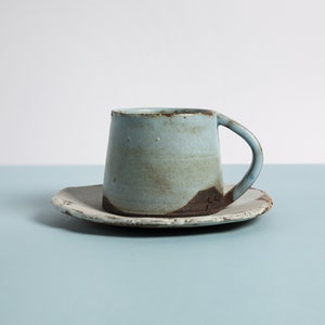 May include: A light blue ceramic coffee cup and saucer with a rustic, hand-thrown look. The cup has a slightly flared rim and a handle. The saucer has a slightly irregular shape.