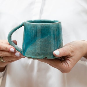 Puede incluir: Una taza de cerámica azul turquesa con una forma única y ligeramente irregular. La taza tiene una base ancha y redondeada y un borde ligeramente acampanado. El asa está unida al lado de la taza y se curva hacia arriba para encontrarse con el borde.