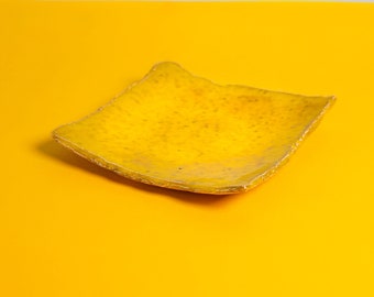 May include: A square, yellow ceramic plate with a textured surface and irregular edges. The plate is set against a bright yellow background with a gray upper section.