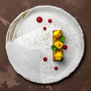 May include: A white plate with a small, rectangular piece of food topped with yellow flowers and green leaves. The food is surrounded by small red dots and a light dusting of brown specks.