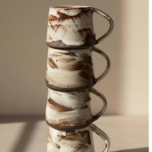 May include: Four ceramic mugs stacked. Each mug features a white base with brown brushstroke patterns, a dark brown rim, and a handle. The mugs are arranged vertically.