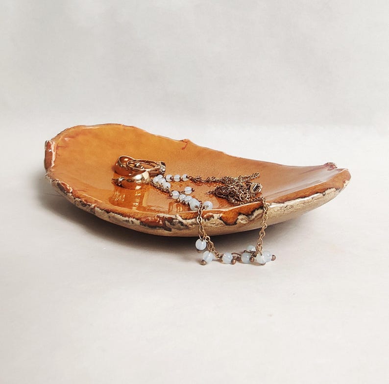 Puede incluir: Un plato de cer&aacute;mica con un esmalte marr&oacute;n y un borde rugoso y texturizado. El plato est&aacute; lleno de joyas, incluyendo un collar de cadena de oro y un par de aretes de aro de oro.