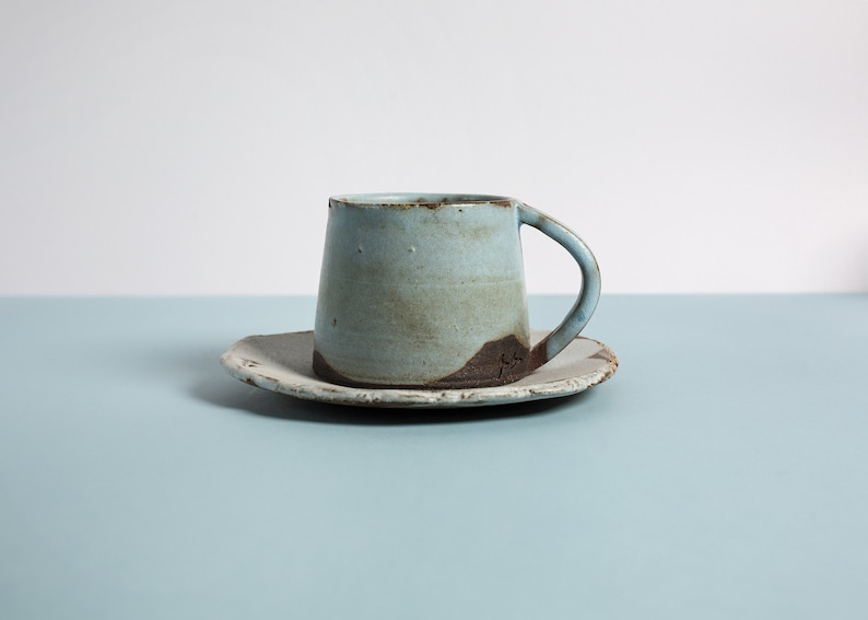 May include: A light blue ceramic coffee cup and saucer with a rustic, hand-thrown look. The cup has a slightly flared rim and a handle. The saucer has a slightly irregular shape.