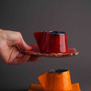 May include: Two handmade ceramic coffee cups with matching saucers. The top cup is red with a blue rim and the bottom cup is orange with a blue rim.