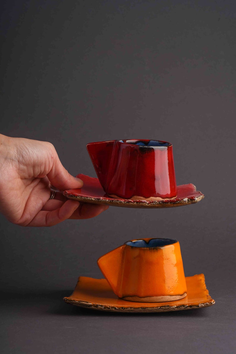 May include: Two handmade ceramic coffee cups with matching saucers. The top cup is red with a blue rim and the bottom cup is orange with a blue rim.