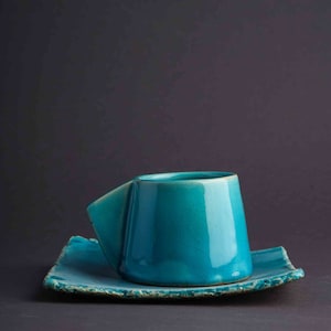 May include: A teal blue ceramic teacup and saucer set. The cup has a unique, angular design and the saucer has a slightly rough, textured edge.