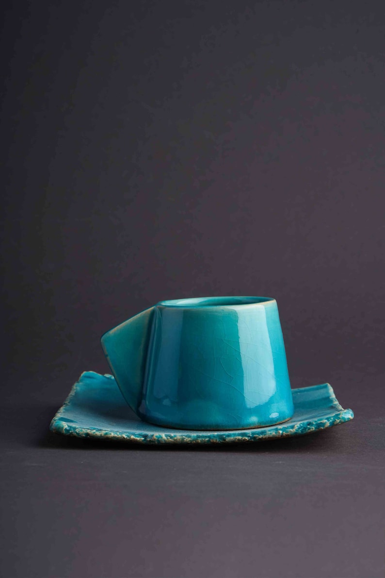 May include: A teal blue ceramic teacup and saucer set. The cup has a unique, angular design and the saucer has a slightly rough, textured edge.