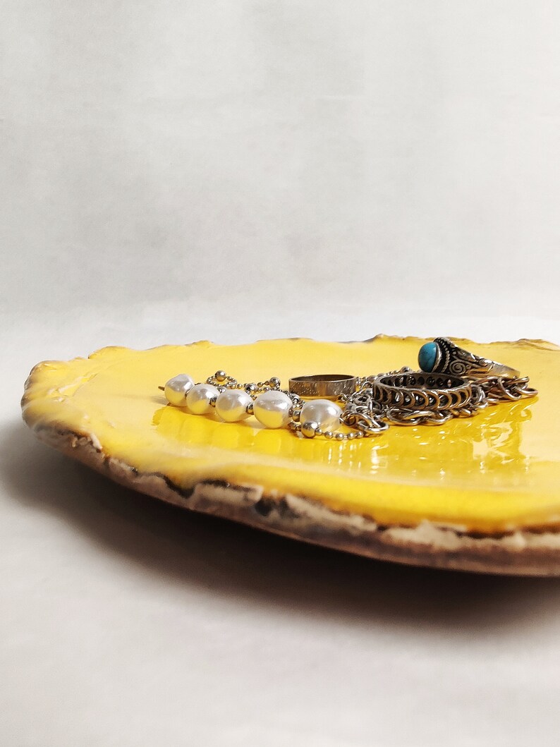 Puede incluir: Un plato de cer&aacute;mica amarillo con un borde rugoso y texturizado. El plato contiene un collar de cadena de plata con cuentas de perlas blancas, un anillo de plata y un anillo de plata con una piedra turquesa.