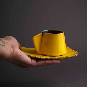 May include: A yellow ceramic coffee cup and saucer with a blue rim. The cup has a unique, asymmetrical shape.
