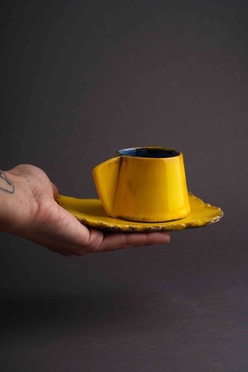 May include: A yellow ceramic coffee cup and saucer with a blue rim. The cup has a unique, asymmetrical shape.